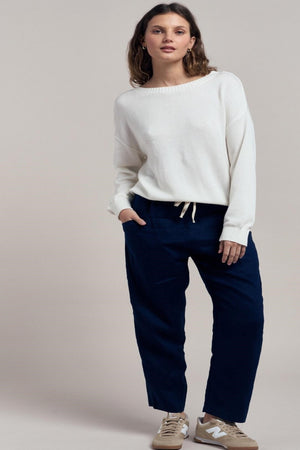 Woman wearing a white sweater and navy pants on a plain background