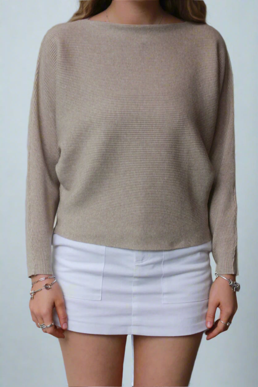 A beige knit sweater worn over a white skirt by a woman, with her arms at her sides and a neutral expression on her face.