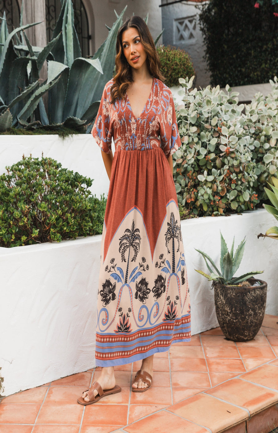 Woman in a patterned dress standing outdoors with plants and a white wall in the background