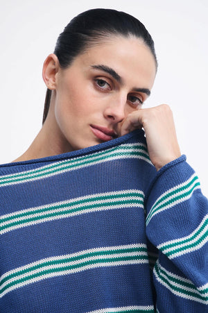 A close-up of a woman wearing a blue knit sweater with horizontal white and green stripes, resting her chin on her hand against a plain light background.